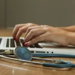 Clinician typing notes on a laptop with a stethoscope nearby